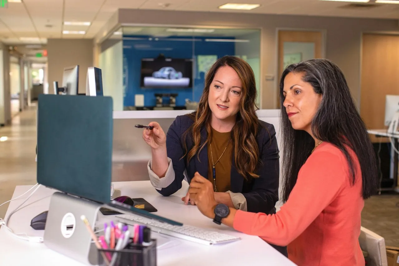 A person sitting at a desk, smiling and working on a laptop with a cup of coffee nearby, in a modern office setting. The image conveys a positive and productive work environment.
