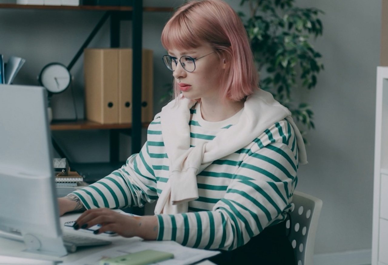 Giovane donna dai capelli rosa e occhiali che digita al computer su una scrivania in un ufficio moderno con scaffali e piante sullo sfondo