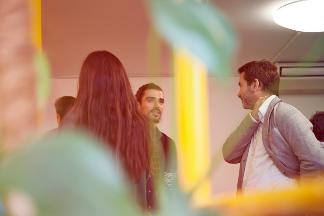 Tre professionisti in abbigliamento smart casual conversano informalmente in un ufficio luminoso con piante sfocate in primo piano