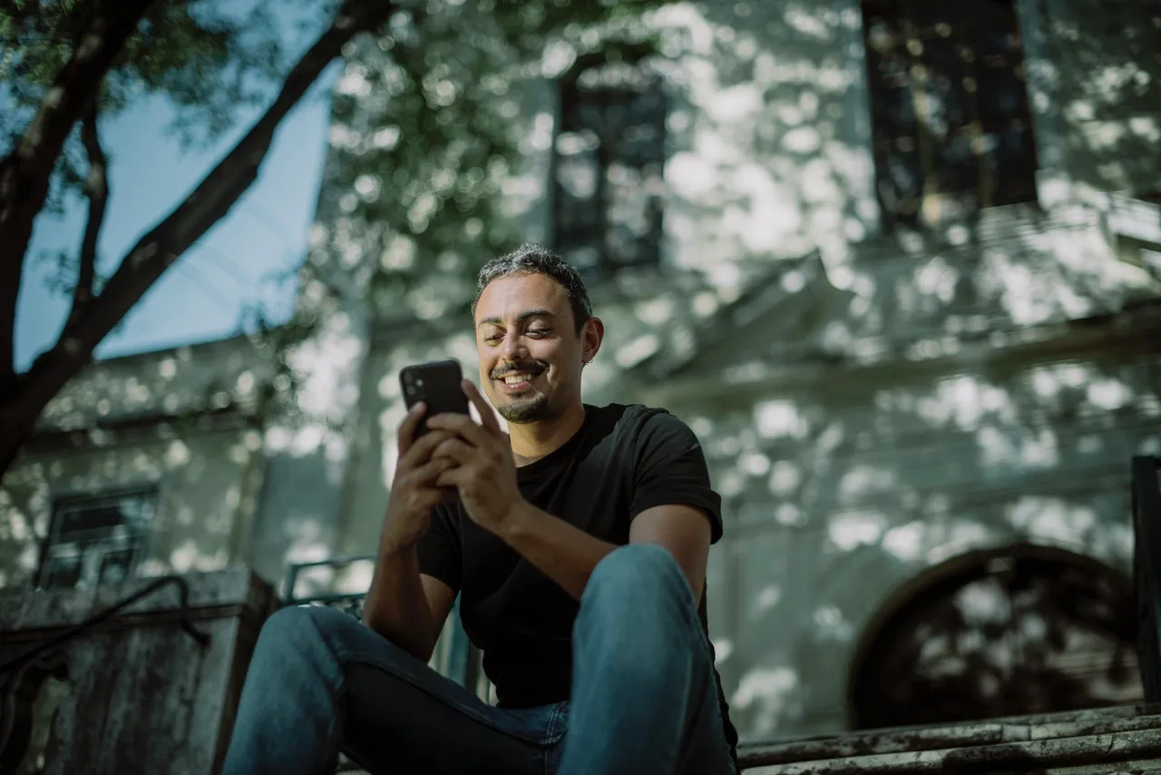 Uomo seduto su gradini allaperto sorride mentre consulta il suo smartphone con edifici storici e alberi sullo sfondo