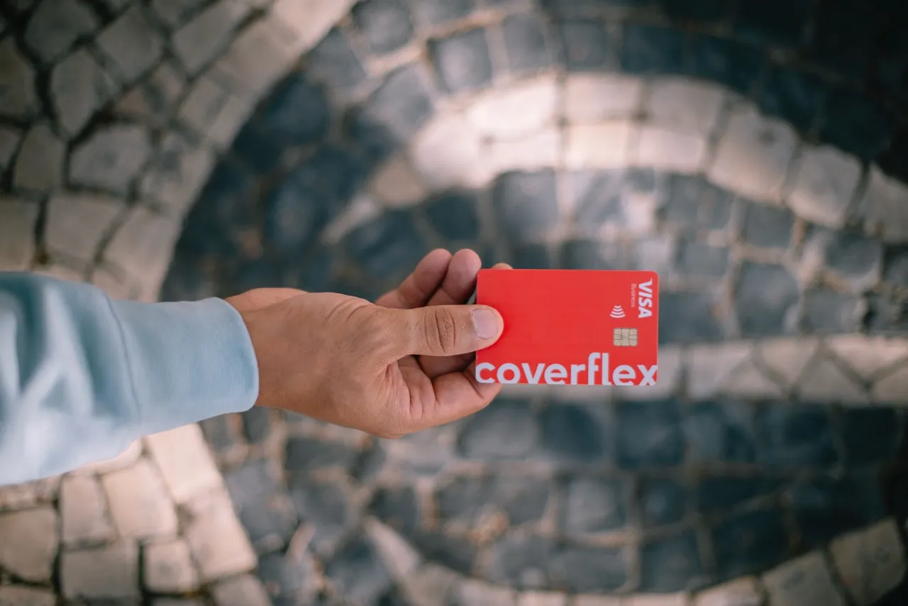 Una mano in camicia azzurra mostra una Coverflex Voucher Card rossa con chip Visa e simbolo contactless davanti a un pavimento di pietre bianche e scure disposte a cerchi
