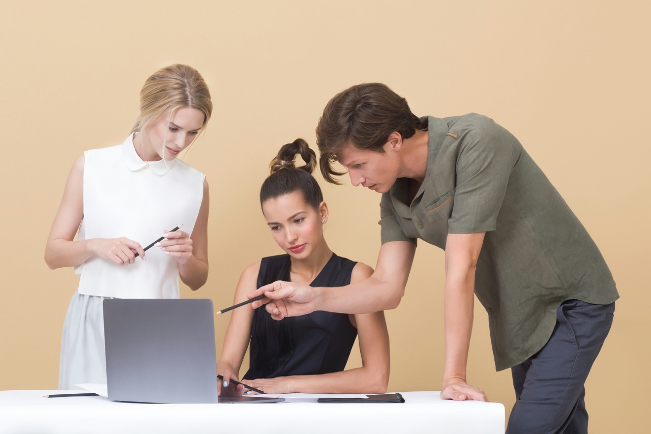 Un uomo indica lo schermo di un laptop mentre due donne, una seduta e l’altra in piedi, osservano documenti su un tavolo bianco in un ambiente minimalista color crema