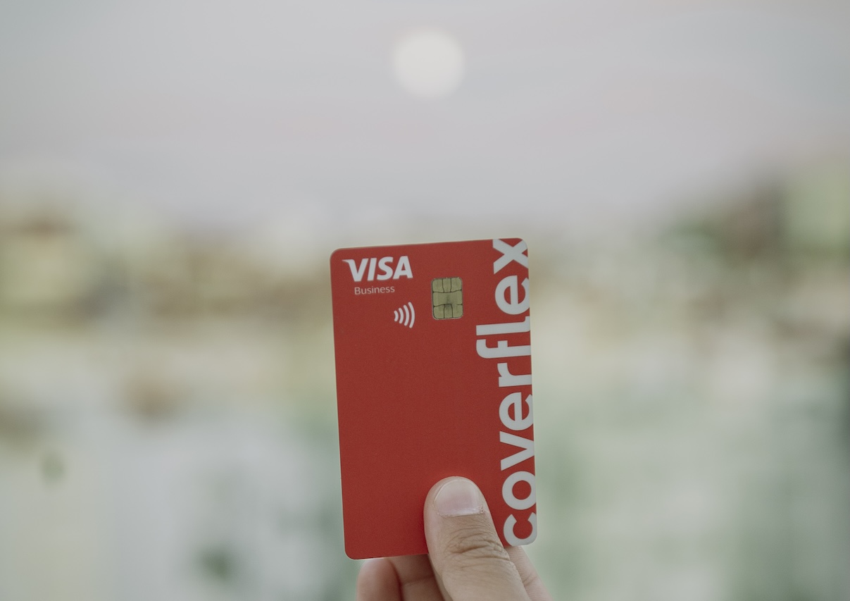 Mano che regge una carta di debito Coverflex rossa con logo Visa Business, chip e simbolo contactless su sfondo cittadino sfocato