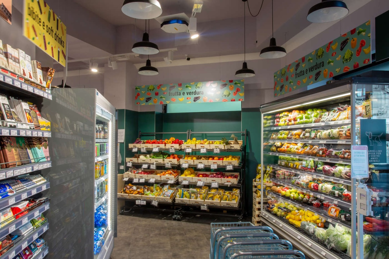 Interno di un negozio to.market in stazione con ceste di frutta e verdura su scaffali, corridoio tra frigoriferi a destra e scaffali di snack a sinistra, carrellini azzurri in primo piano