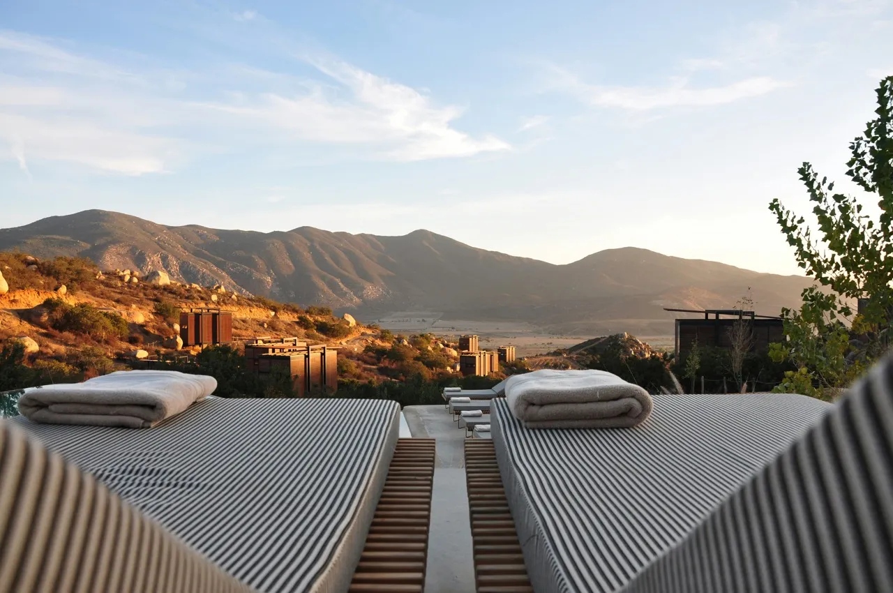 Due lettini con materassini a righe e asciugamani arrotolati su un terrazzo di un resort moderno, con piccole cabine in legno tra rocce e colline panoramiche illuminate dalla luce calda del tramonto