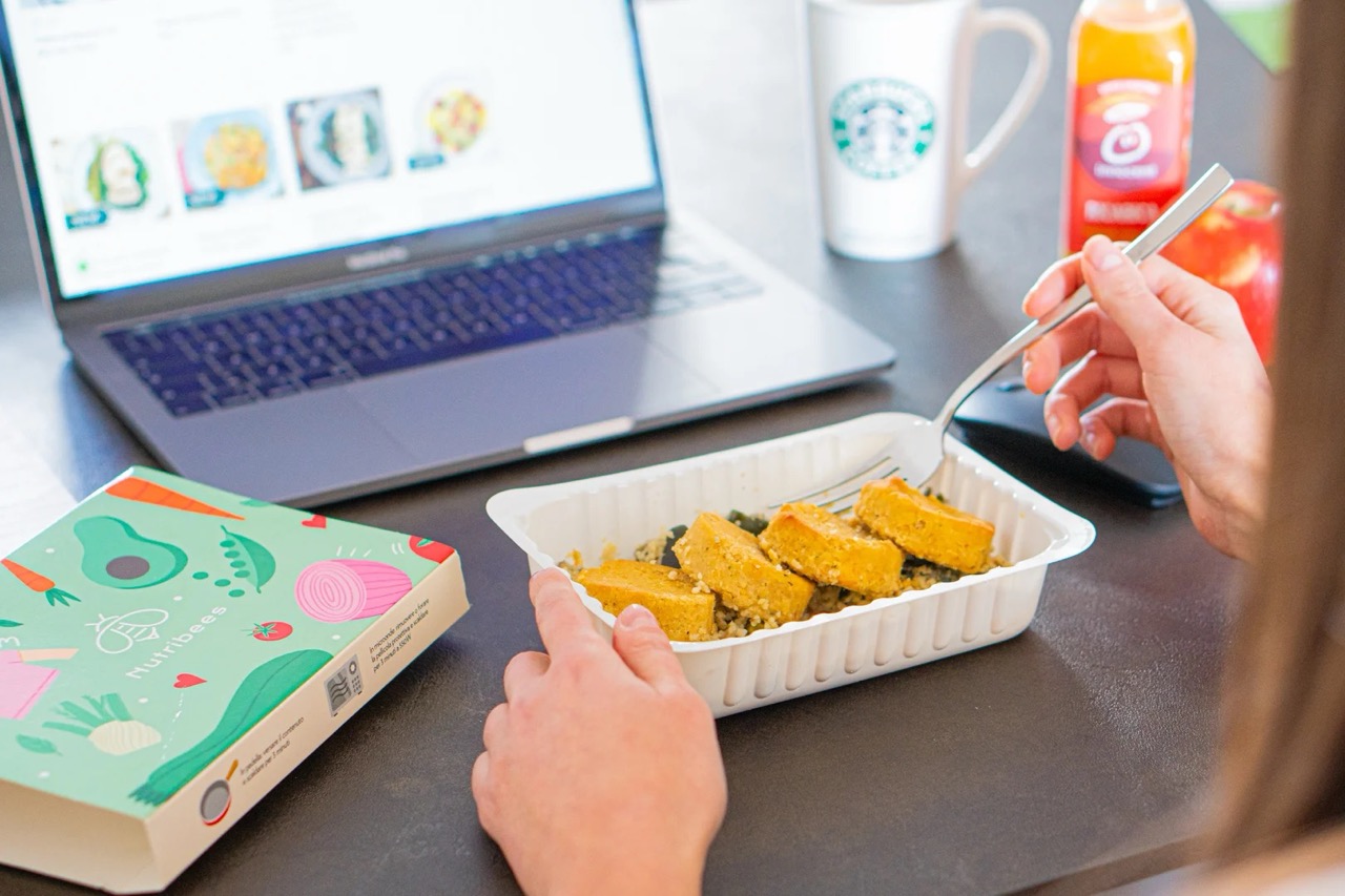 Mano sosteniendo un tenedor mientras come un plato listo de Nutribees en bandeja desechable sobre una mesa con un portátil, taza de café, botella de zumo y manzana