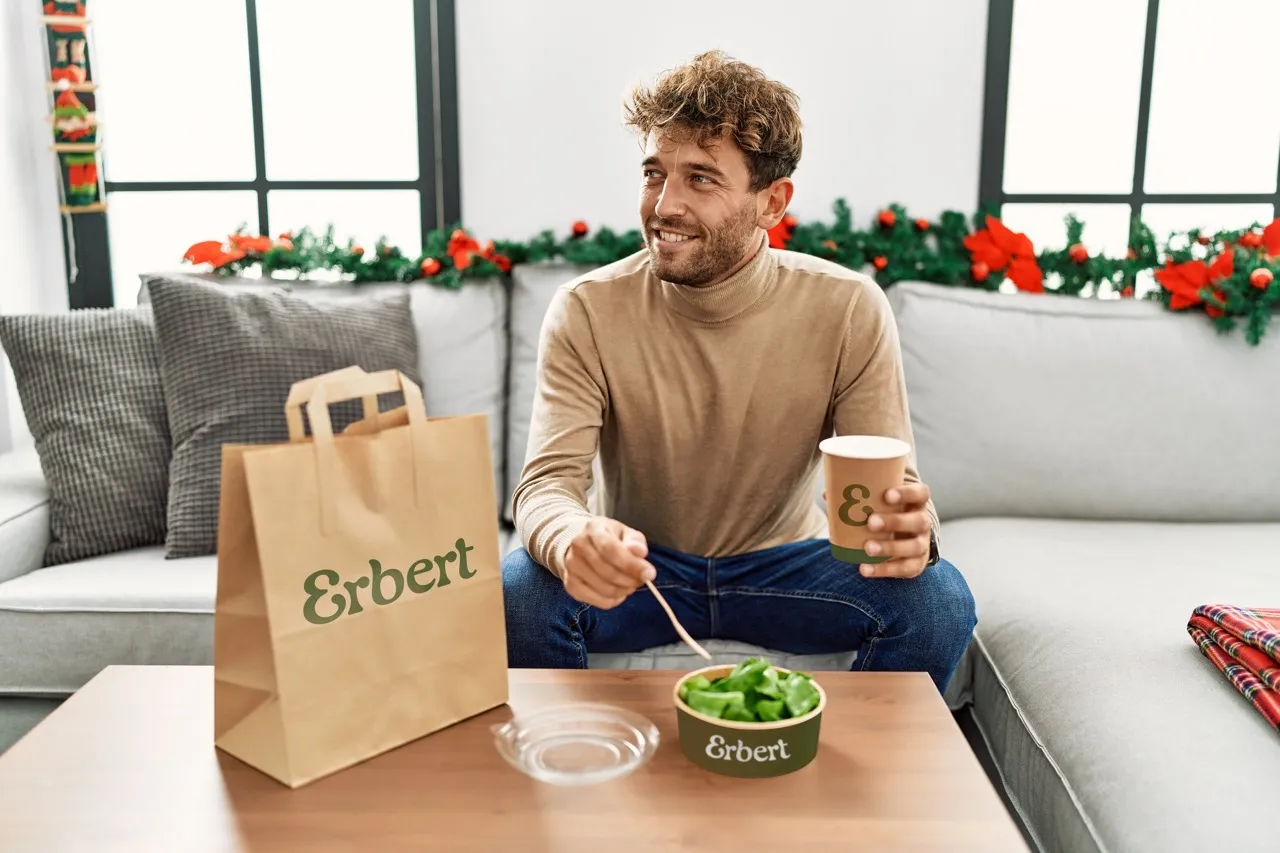 Uomo in abbigliamento casual seduto su un divano con sacchetto di carta Erbert, ciotola di insalata e bicchiere da asporto su un tavolino, con ghirlande e decorazioni natalizie sullo sfondo