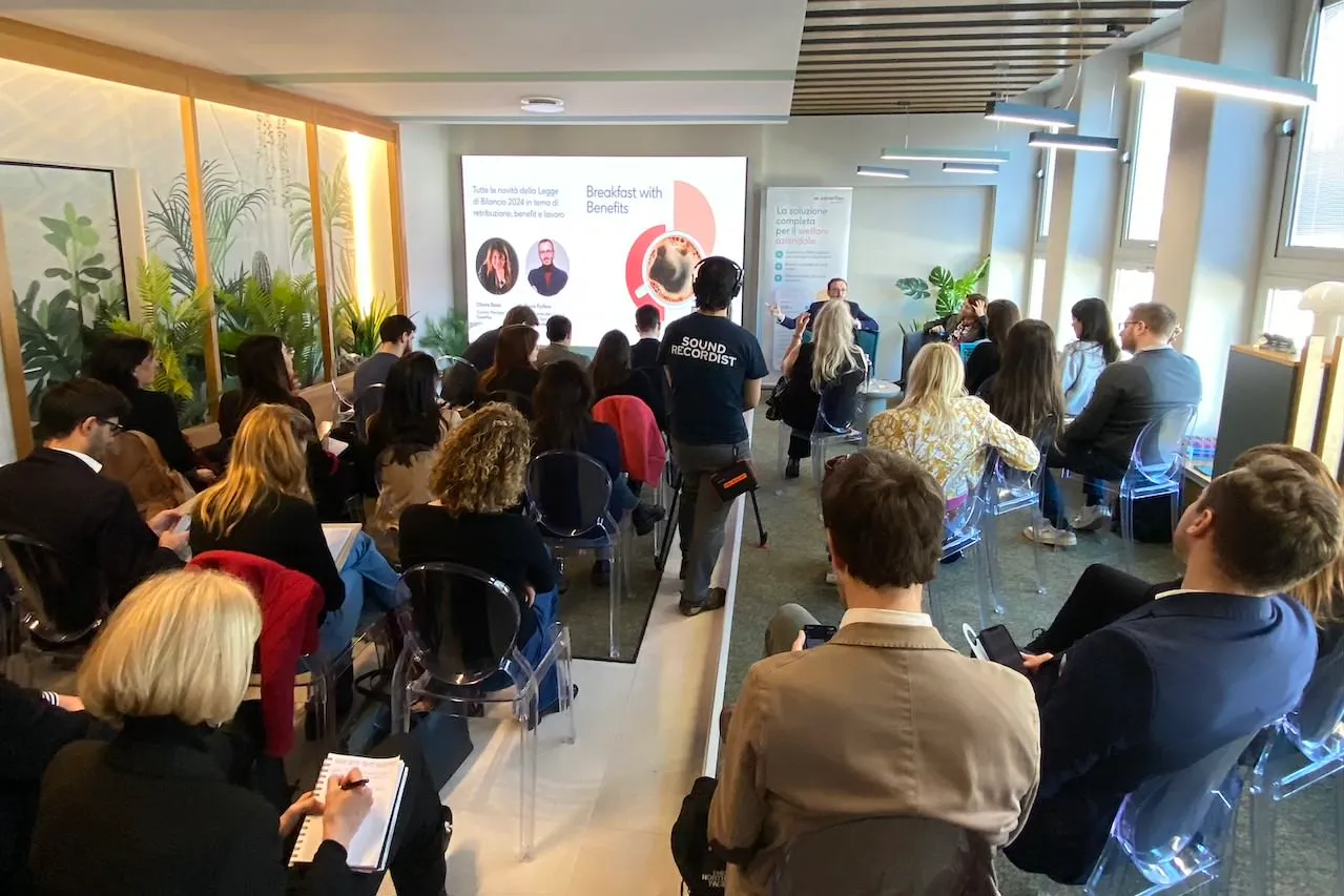 Partecipanti seduti su sedie trasparenti seguono un intervento su benefit aziendali durante l evento Breakfast with Benefits con relatori sul palco e operatore audio con microfono