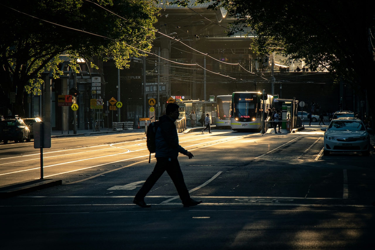 Uomo con zaino attraversa un incrocio urbano al tramonto mentre un autobus si ferma alla fermata evidenziando il tema della mobilita sostenibile