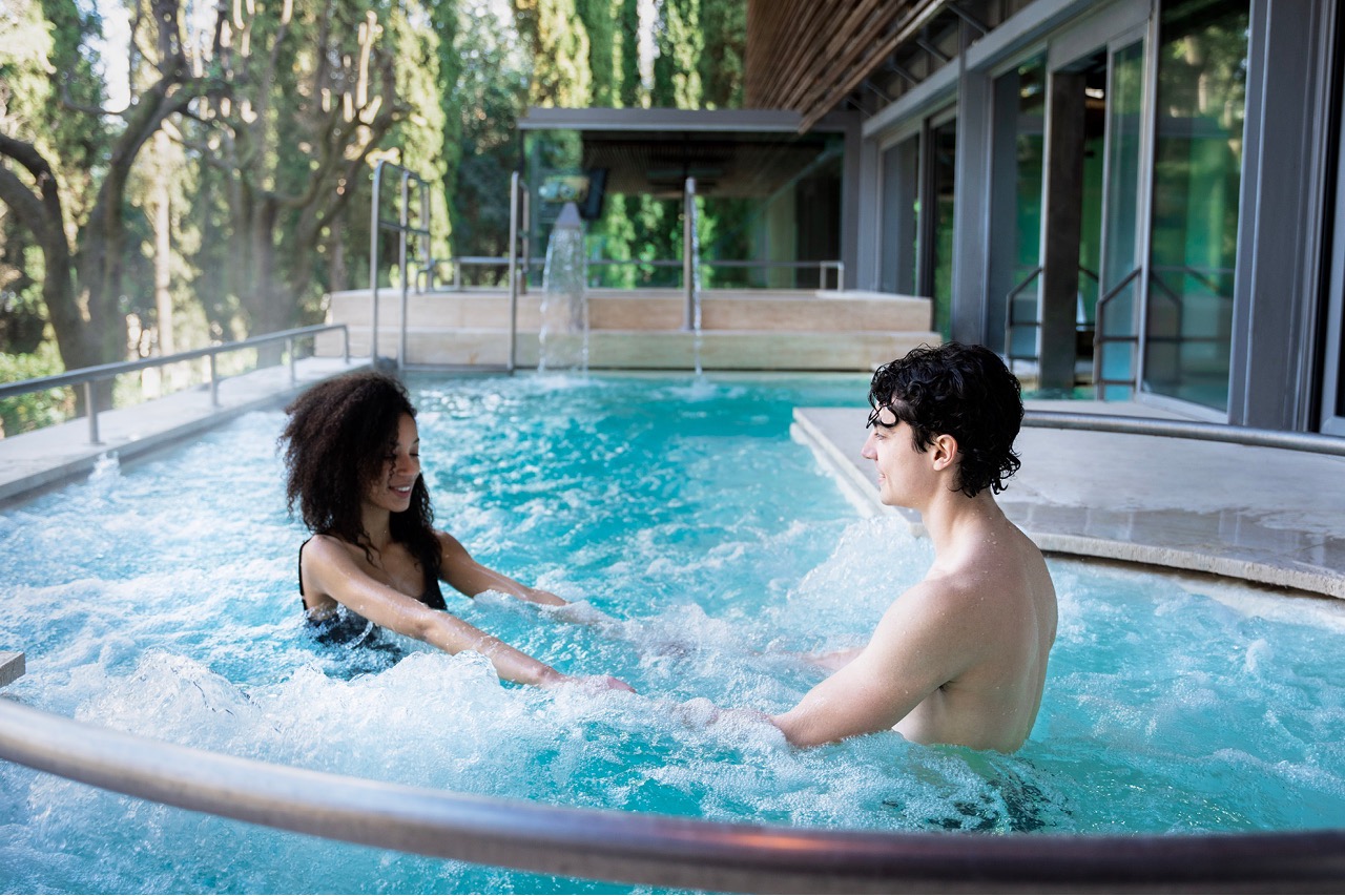 Coppia di giovani in una piscina termale moderna che si tengono per mano tra getti idromassaggio circondati da vegetazione e ampie vetrate