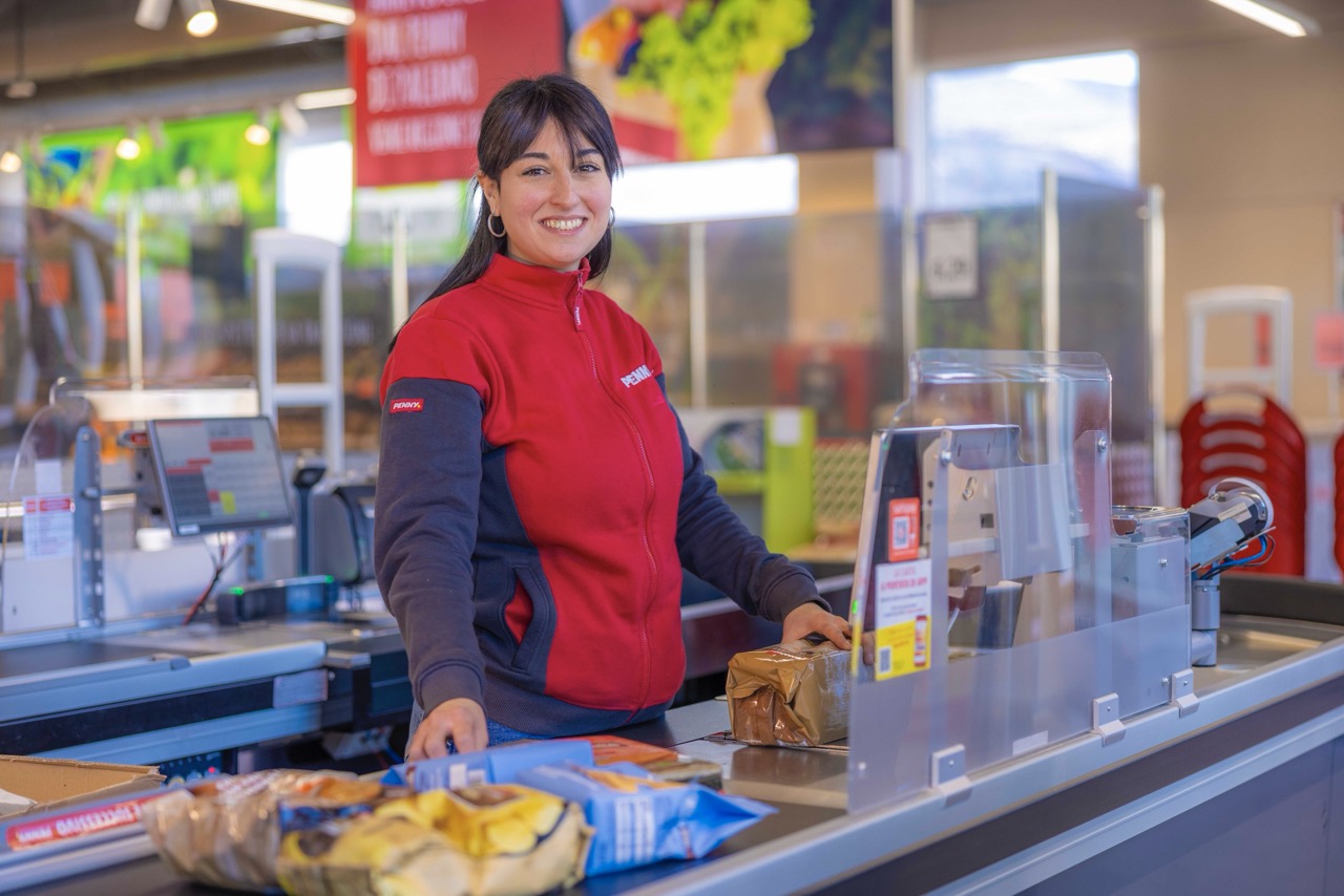 commessa Penny sorridente al banco cassa dietro barriera in plexiglas che scansiona prodotti sul nastro trasportatore
