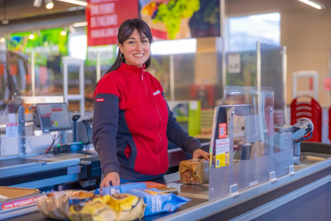 commessa Penny sorridente al banco cassa dietro barriera in plexiglas che scansiona prodotti sul nastro trasportatore