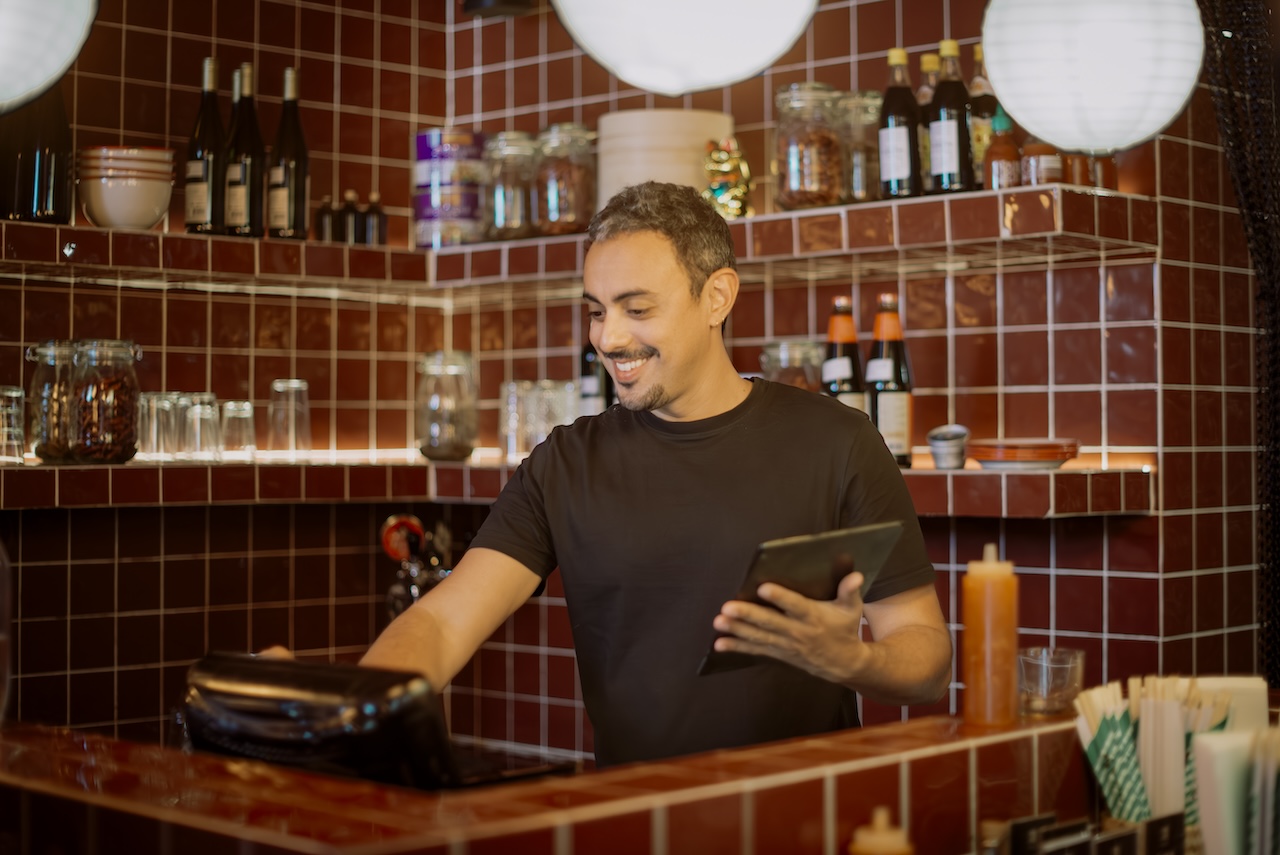 Barista in maglietta nera sorride mentre prende ordini con un tablet e li registra sul registratore di cassa davanti a scaffali con bottiglie, barattoli e stoviglie in un ristorante con rivestimenti in mattonelle rosse e luci sospese