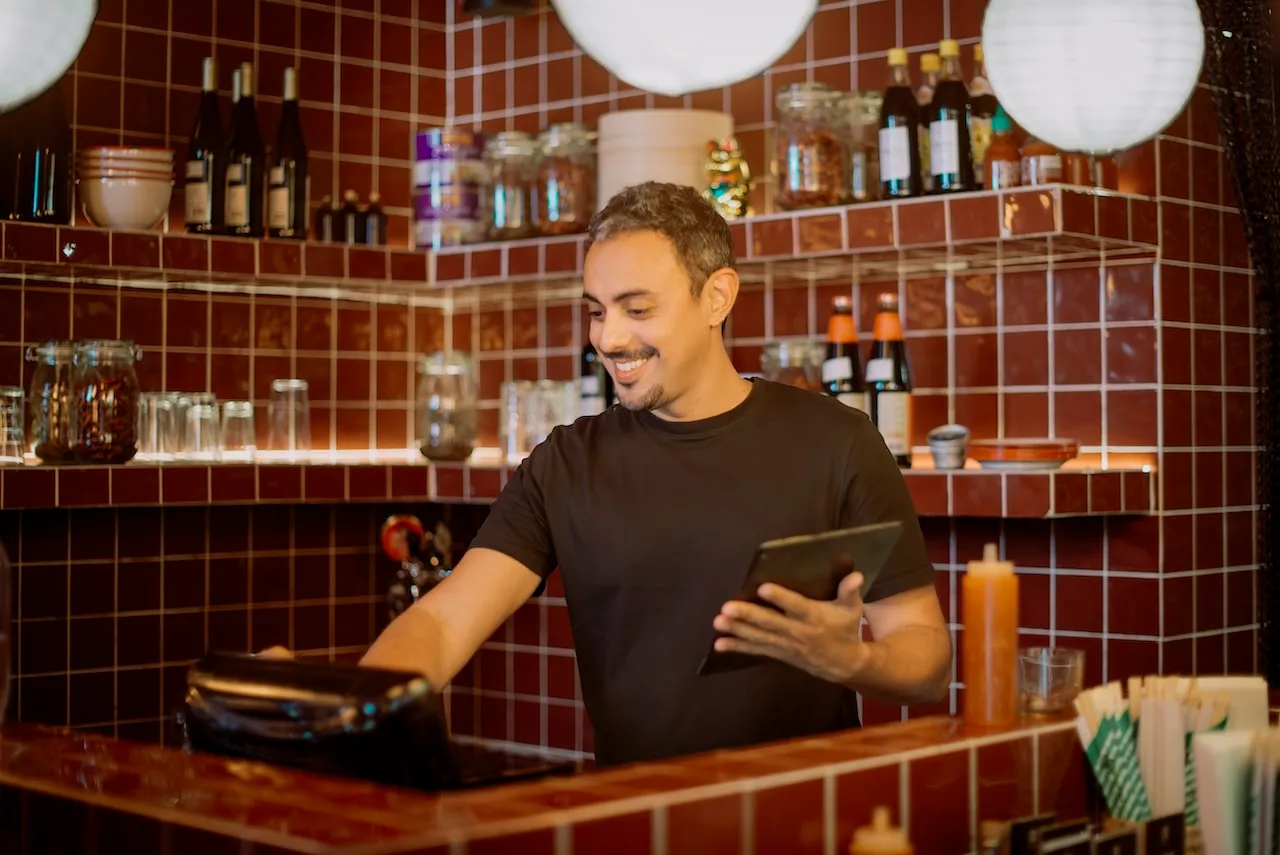 Barista in maglietta nera sorride mentre prende ordini con un tablet e li registra sul registratore di cassa davanti a scaffali con bottiglie, barattoli e stoviglie in un ristorante con rivestimenti in mattonelle rosse e luci sospese