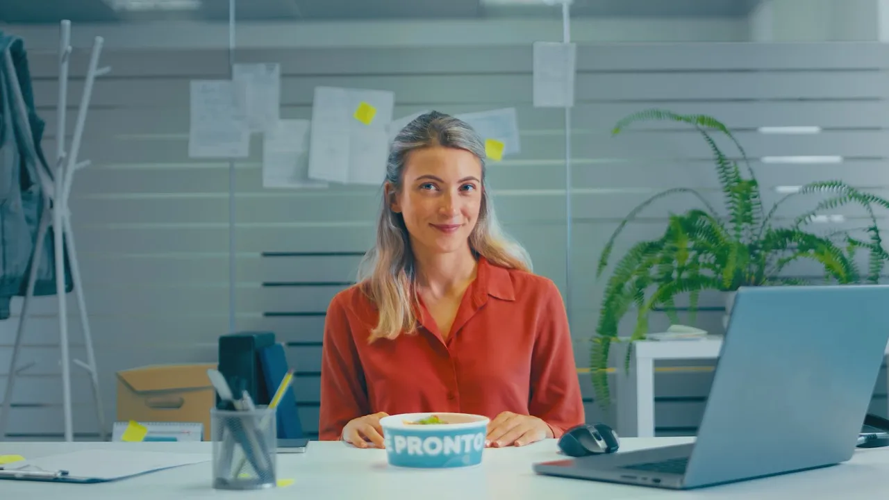 Donna sorridente seduta davanti al computer in un ufficio moderno con un contenitore per il pranzo Pronto sulla scrivania, circondata da pianta, mouse e porta matite