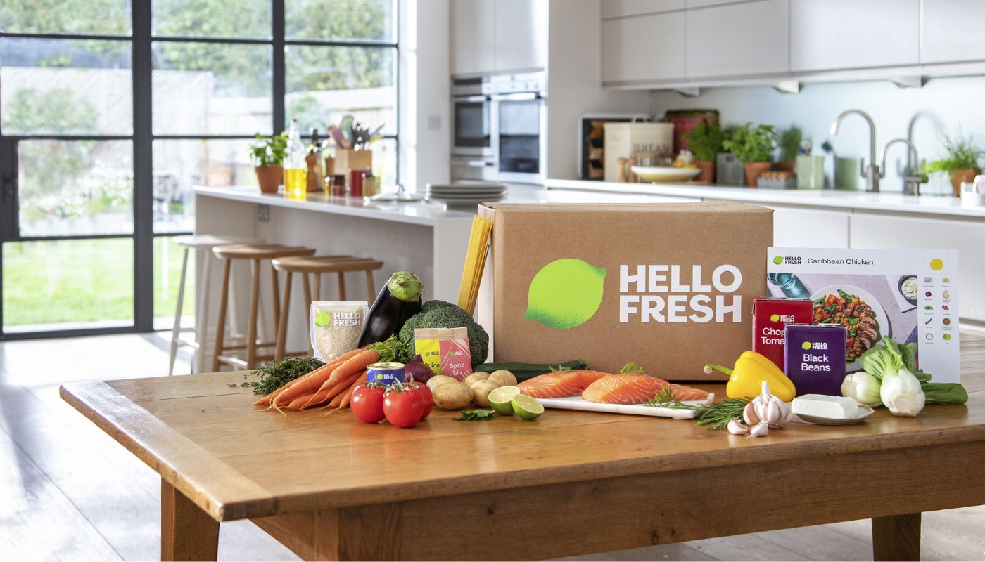 Caja HelloFresh con ingredientes frescos sobre mesa de madera en cocina moderna incluyendo salmón, tomates, zanahorias, brócoli, berenjena, especias, arroz, pasta y tarjeta con receta de pollo caribeño