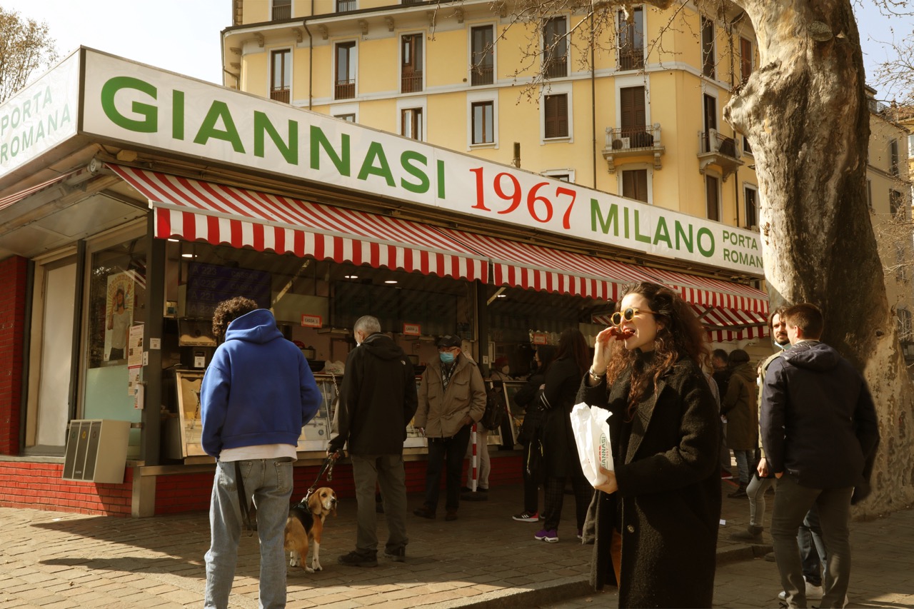 Vista frontale del chiosco Giannasi 1967 Milano Porta Romana con insegna verde e rossa, persone in fila, un cane al guinzaglio e una donna in primo piano che mangia una crocchetta tenendo una busta con logo