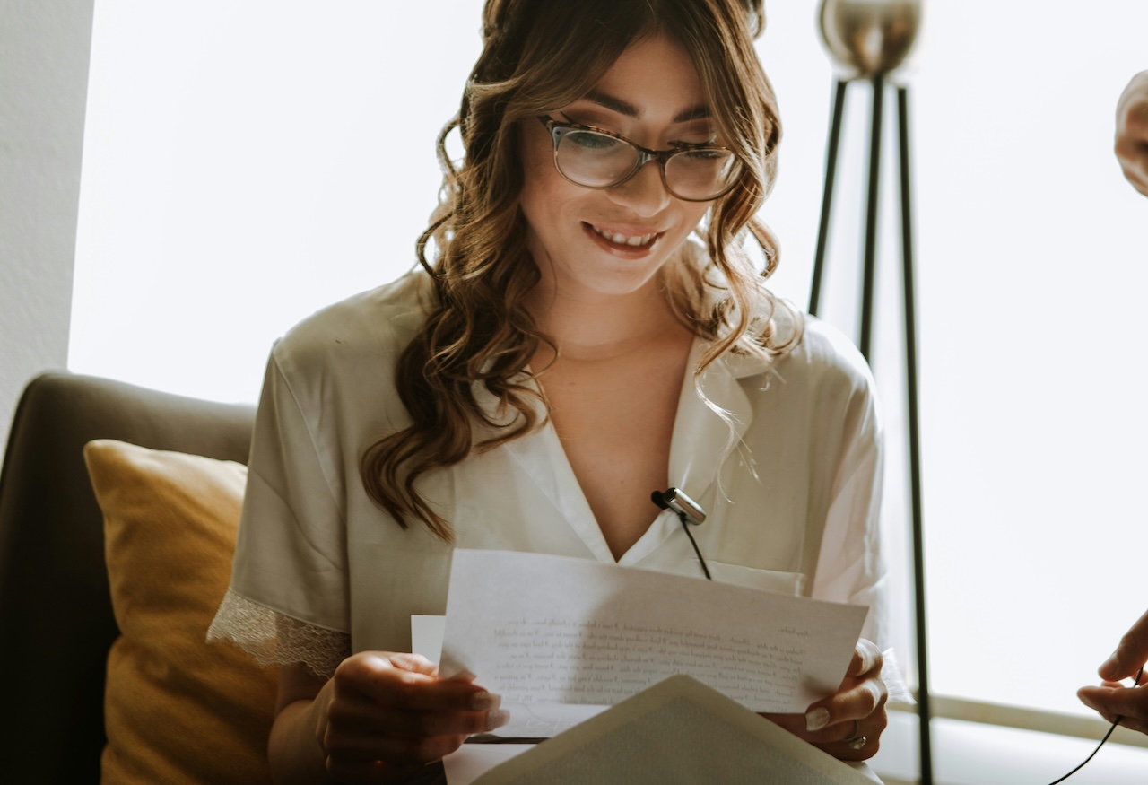 Donna sorridente con occhiali legge una lettera mentre le viene applicato un microfono su una camicia chiara seduta su un divano
