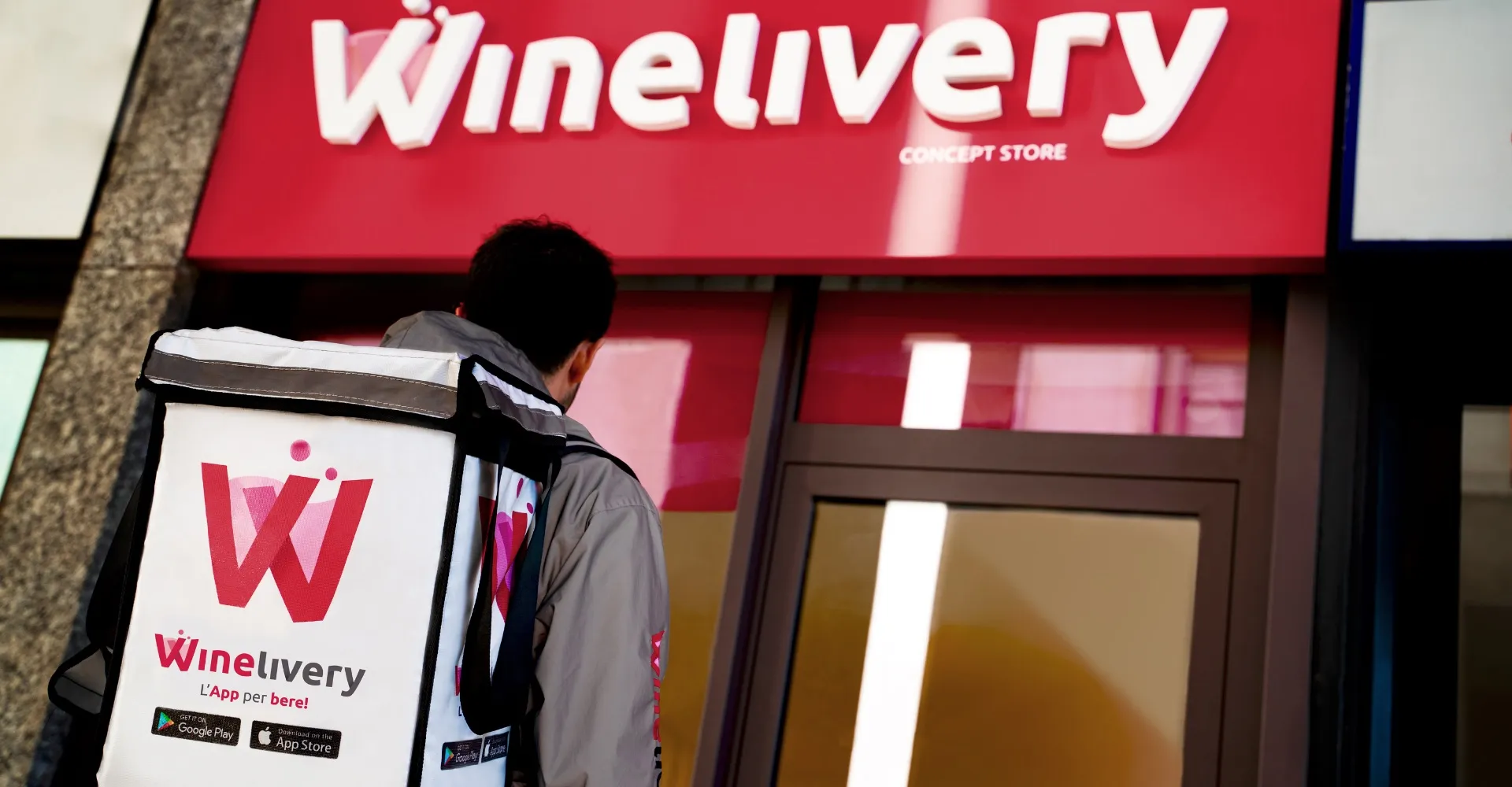 Corriere Winelivery con zaino termico bianco e rosso davanti a vetrina e insegna Winelivery Concept Store