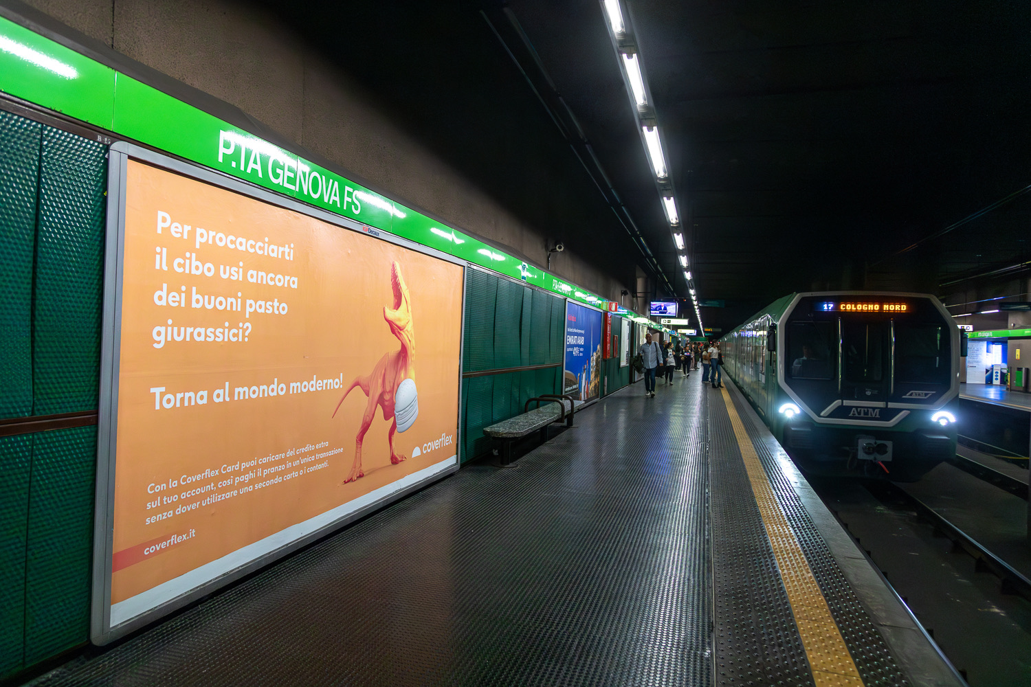 Banchina metro Porta Genova FS con poster arancione Coverflex raffigurante dinosauro e testo su buoni pasto giurassici