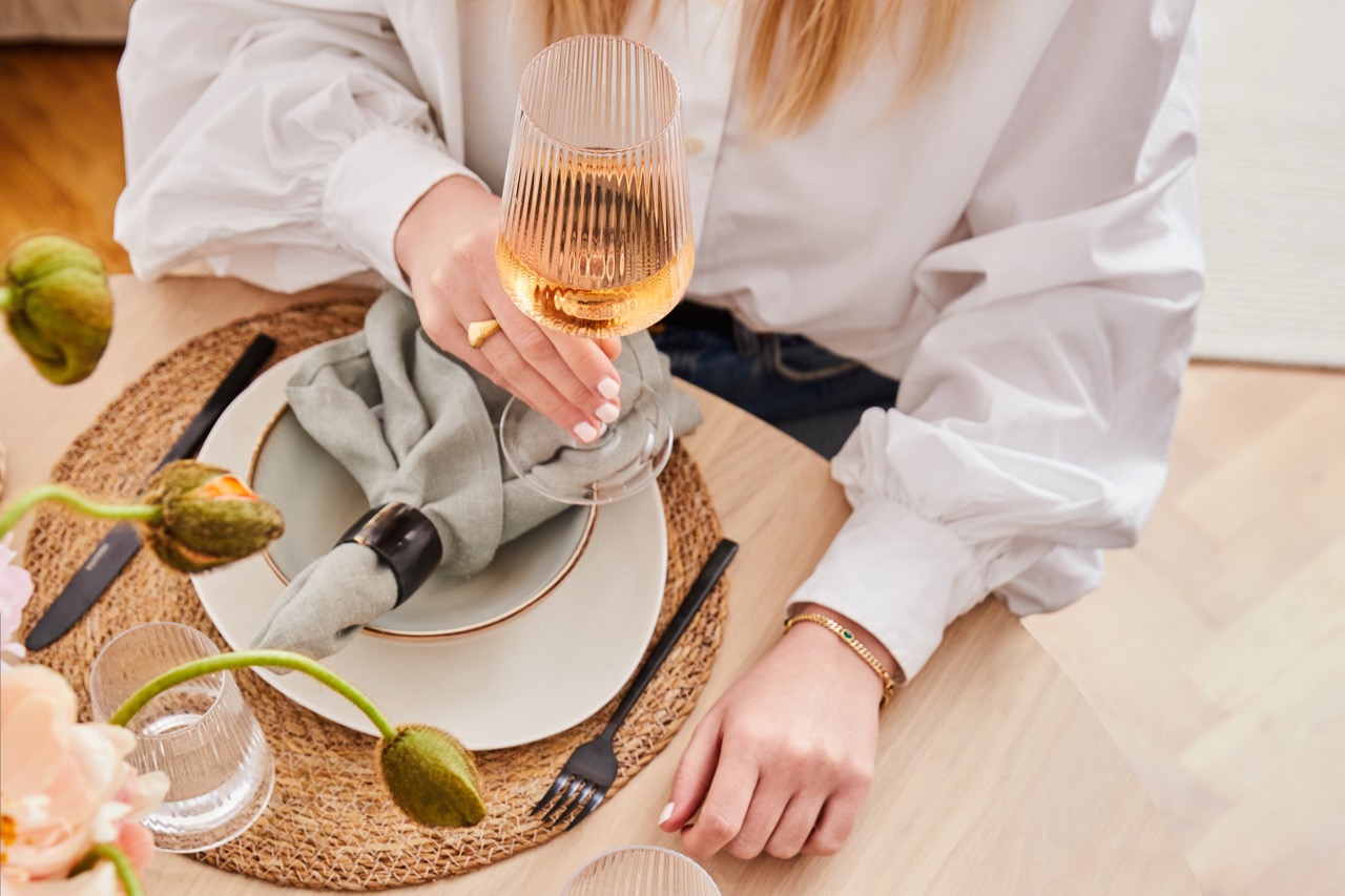 Dettaglio di una tavola da pranzo con piatto su tovaglietta in fibra, tovagliolo fermato da un anello, posate nere, bicchiere d'acqua, fiori in primo piano e mano femminile che impugna un calice di vino rosato