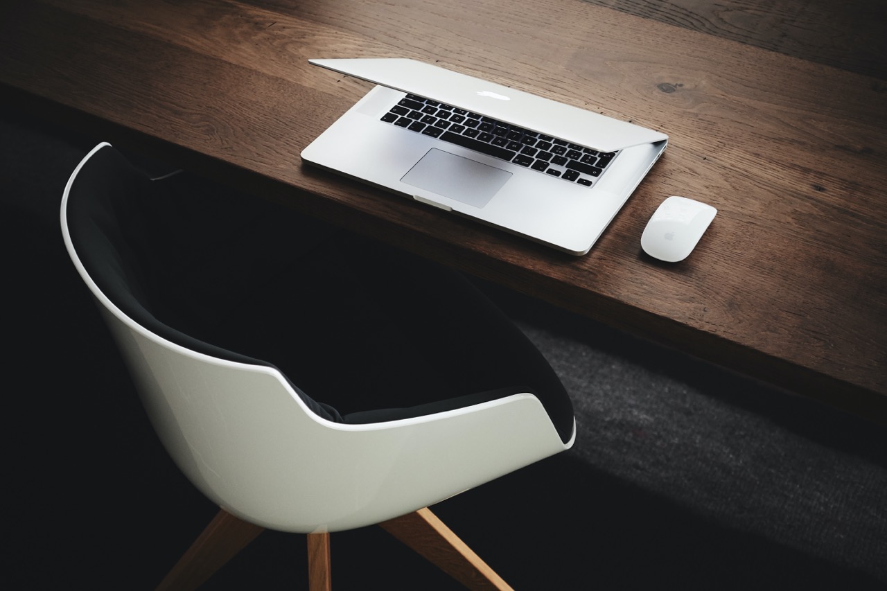 Scrivania in legno scuro con MacBook parzialmente aperto e mouse wireless bianco sul lato accanto a sedia minimalista bianca e nera con gambe in legno