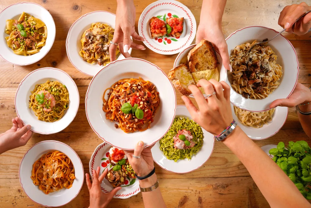Vista aerea di otto piatti di pasta fresca con vari condimenti e bruschette su un tavolo di legno mani di commensali che servono fette di pane