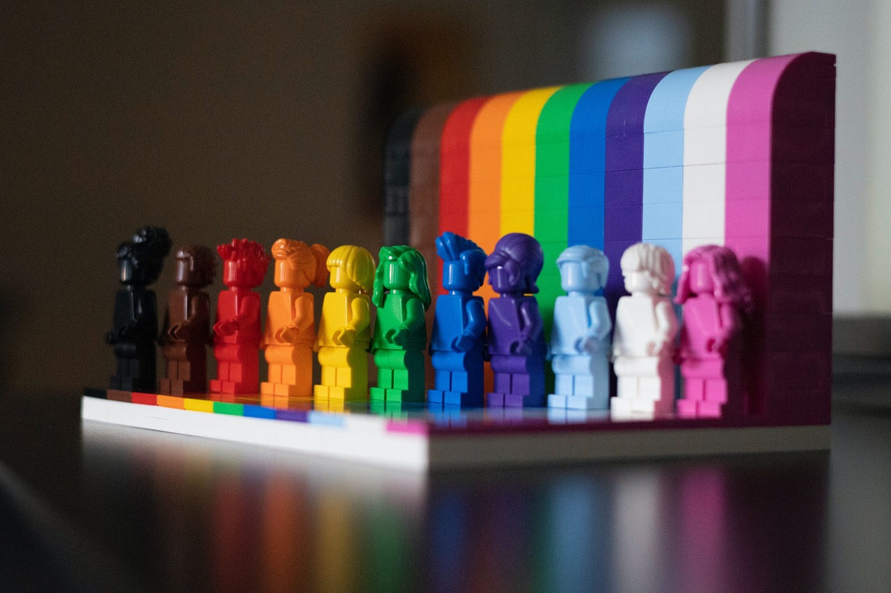 Doce minifiguras de LEGO alineadas sobre una base blanca con bloques verticales detrás en una gradación de 12 colores (negro, marrón, rojo, naranja, amarillo, verde, azul, morado, celeste, blanco, rosa y fucsia) representando la diversidad LGBTQIA+