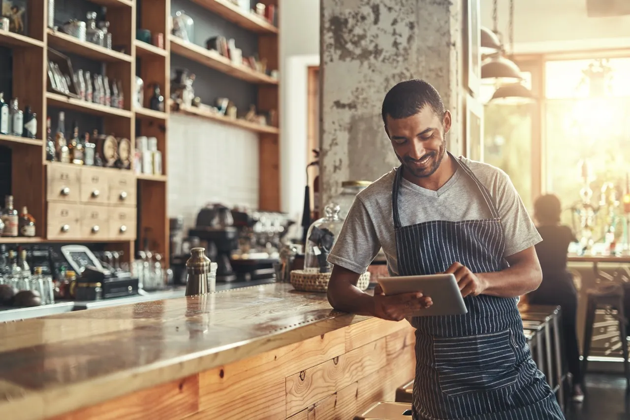 barista sorridente con grembiule a righe che consulta un tablet davanti al bancone di un bar moderno con scaffali di bottiglie sullo sfondo