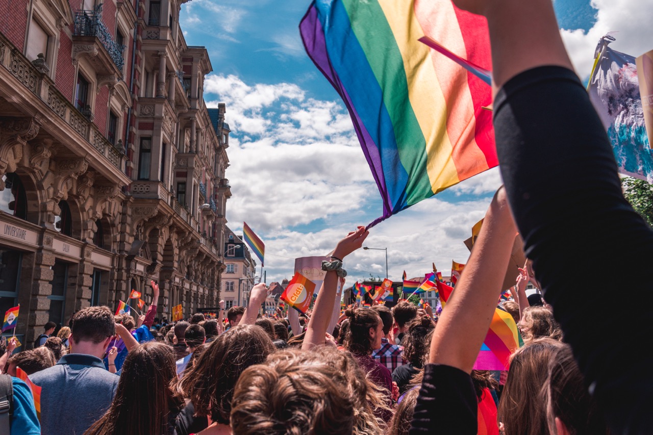 Partecipanti a un Pride in strada con bandiere arcobaleno alzate e un grande stendardo che sventola davanti a un palazzo storico sotto un cielo sereno