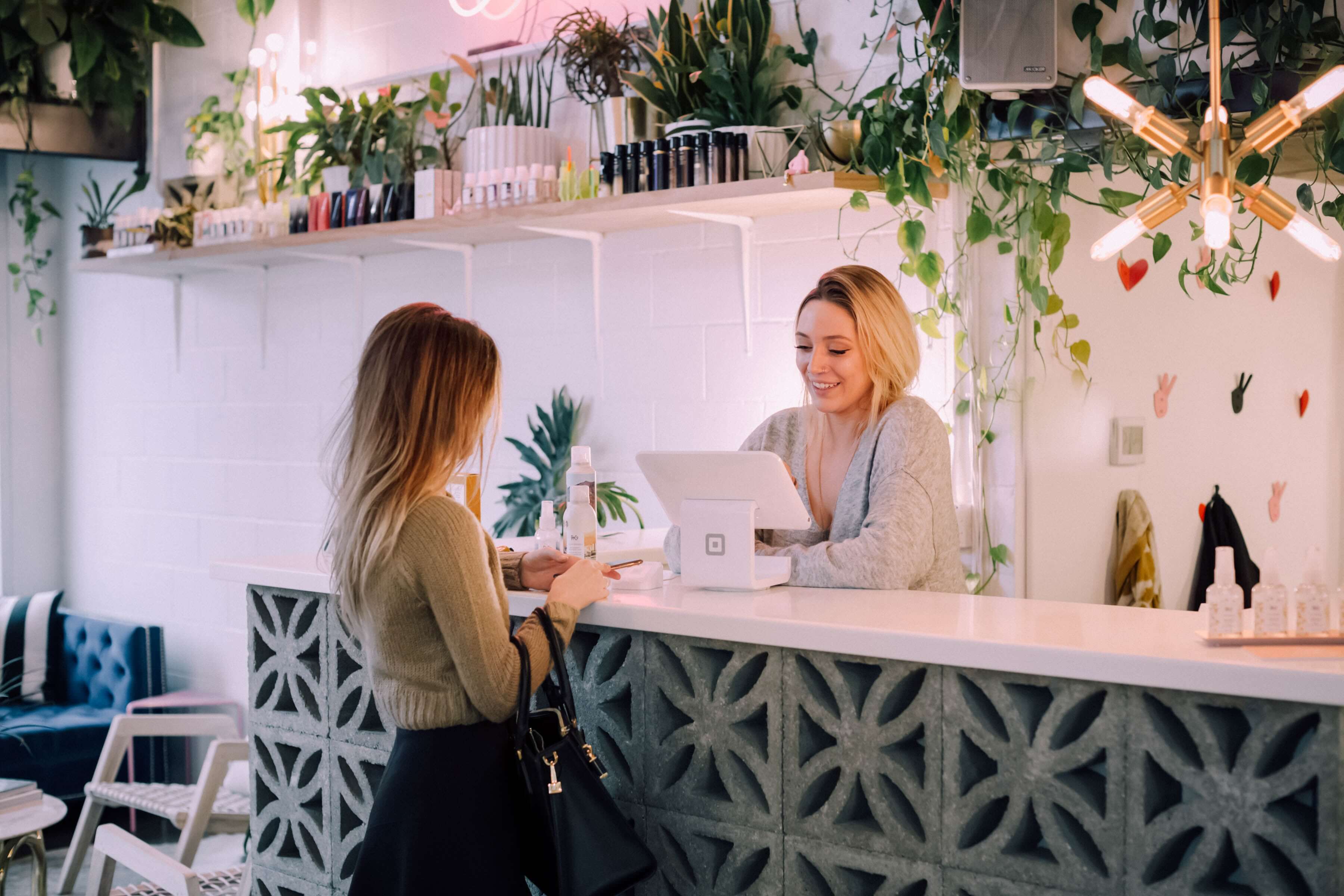 Mujer entrega pago en un mostrador decorado con celosías de hormigón, terminal digital y plantas colgantes, atendida por una cajera sonriente en un local luminoso y moderno