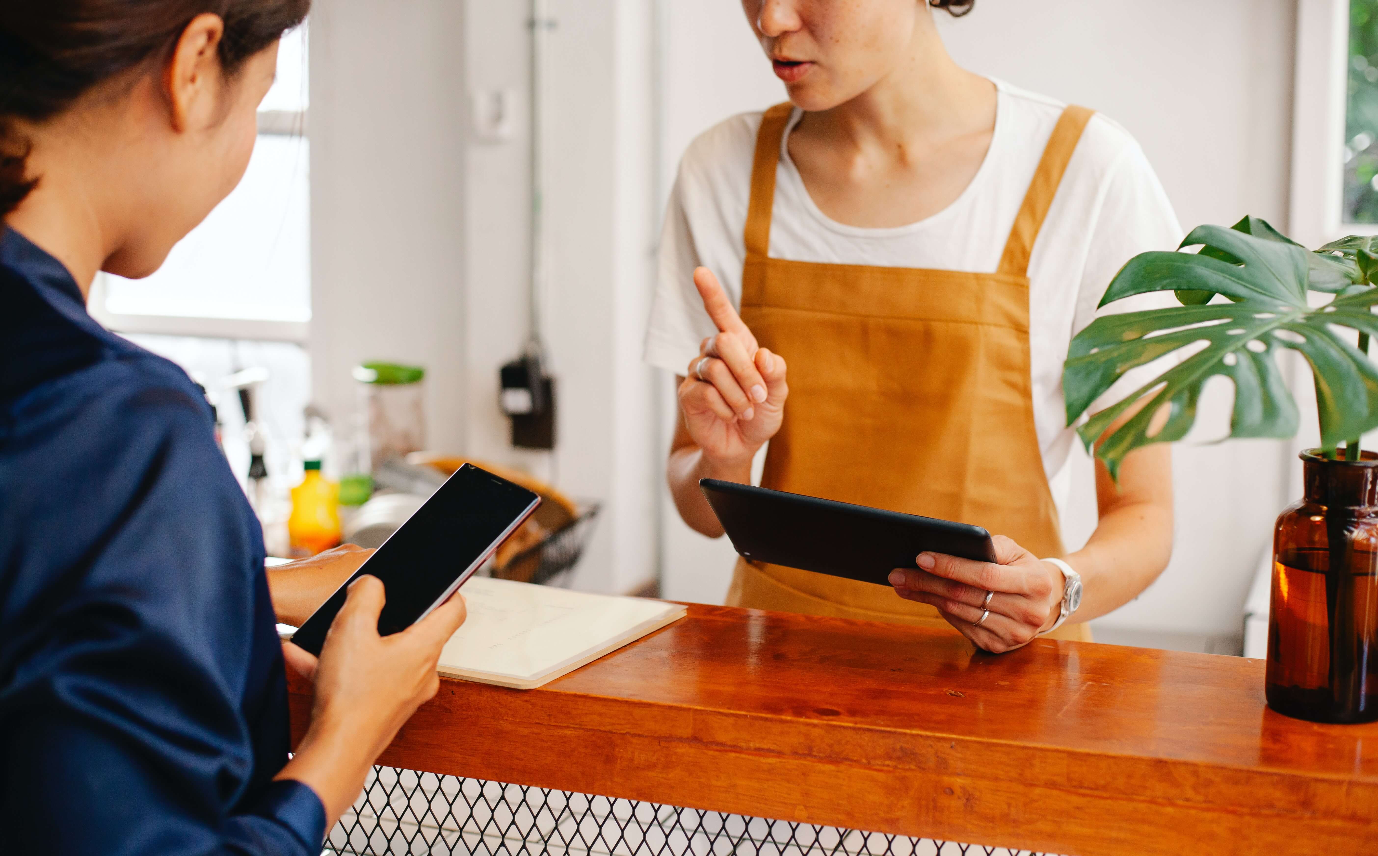 Commerciante in grembiule ocra utilizza un tablet al bancone mentre la cliente mostra lo smartphone