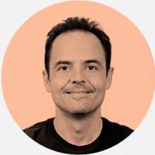 Um homem de cabelos castanhos e pele clara, usando uma camisa azul, está posando em um ambiente externo. O fundo apresenta uma vegetação verde, sugerindo um dia ensolarado e calmo.