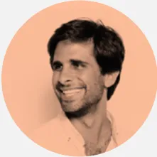 Um homem com camisa azul sorrindo e segurando um copo, em um ambiente iluminado e alegre, que sugere um momento de celebração ou descontração.