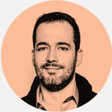 Um homem sorridente usa uma camisa azul e óculos em um ambiente interno com decoração moderna. O fundo apresenta uma estante com prateleiras e objetos decorativos.