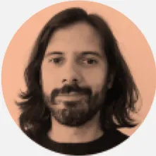 Um homem de óculos e barba, usando uma camisa branca, está sorrindo enquanto segura uma bebida em um copo. Ao fundo, há um cenário de festa com luzes e decorações.