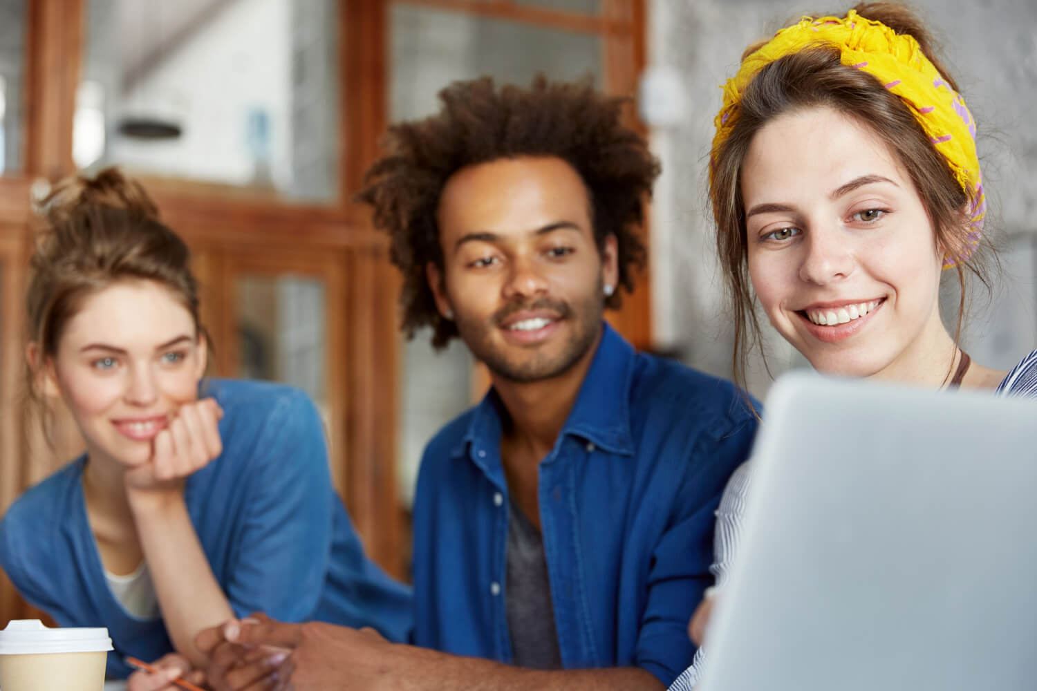 Duas mulheres e um homem de aspecto descontraído colaboram num projeto, focados no ecrã de um portátil sobre a mesa; um copo de café aparece em primeiro plano