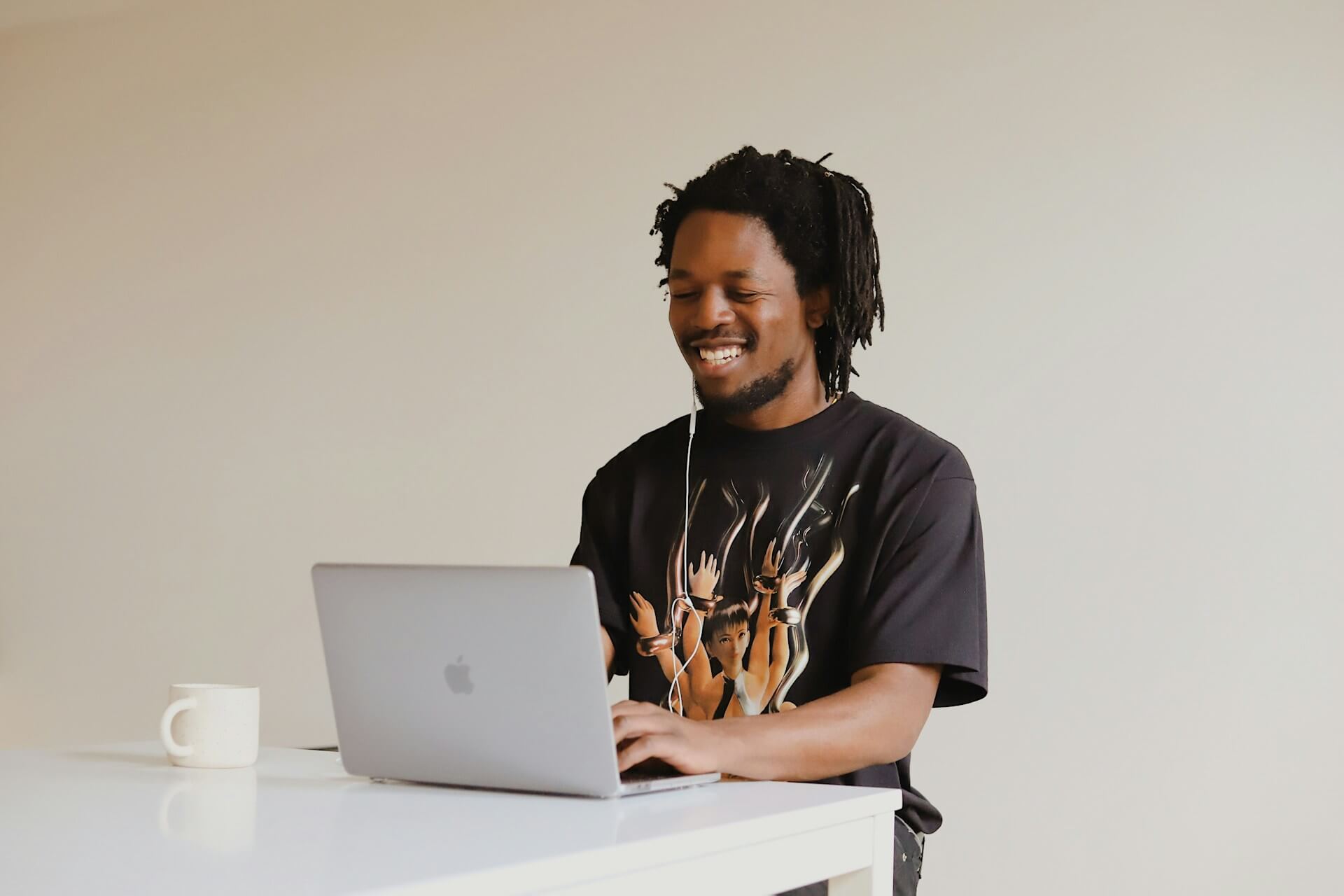 Homem jovem com dreadlocks e auriculares sorri enquanto digita num MacBook sobre mesa branca minimalista, veste t-shirt preta com grafismo de mãos entrelaçadas, caneca de cerâmica ao lado, fundo neutro