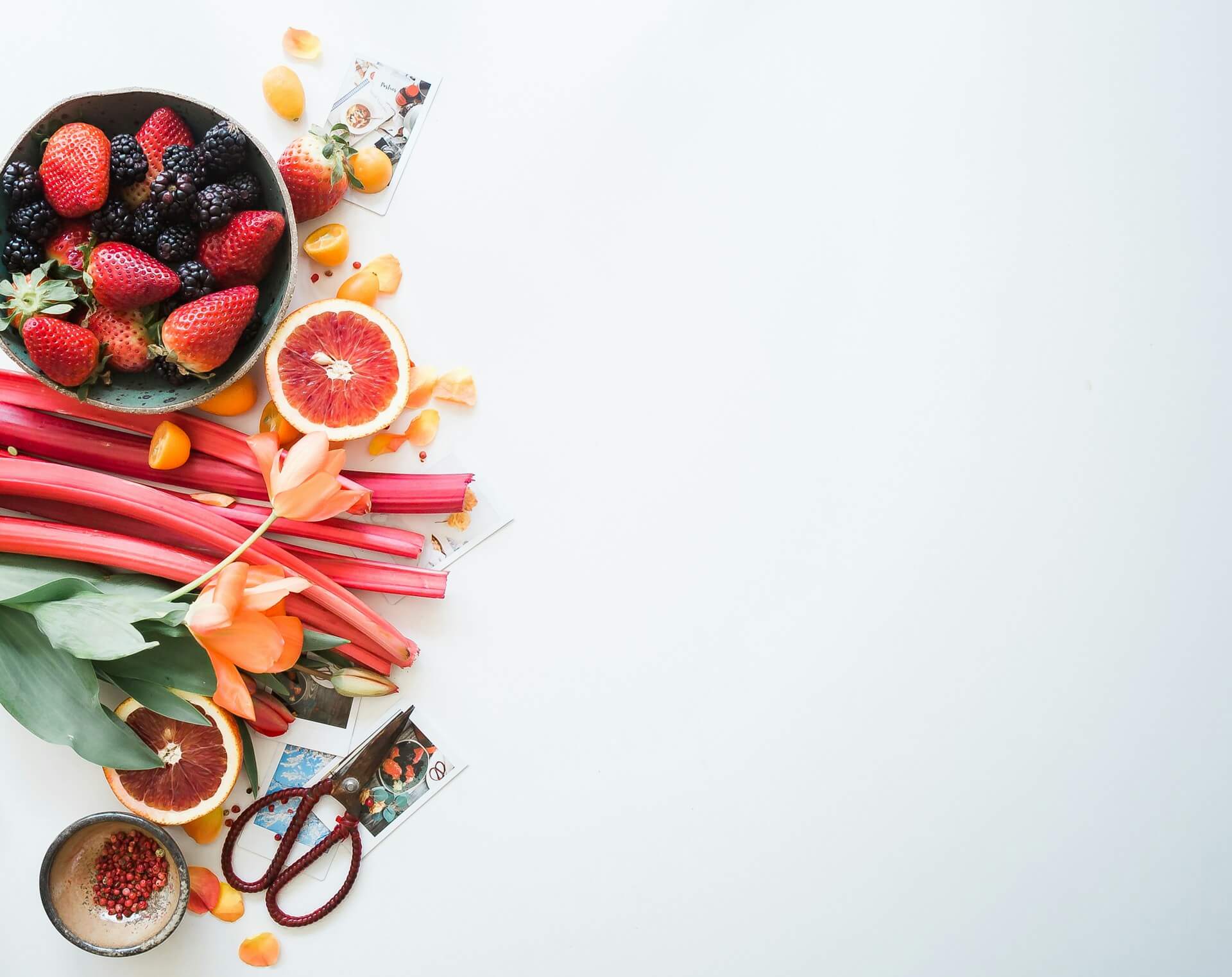 Composição de culinária com tigela de morangos e amoras, metades de laranja sanguínea, pequenas kumquats, talos de ruibarbo, tulipas laranja, sementes de pimenta rosa e tesoura sobre fundo branco minimalista