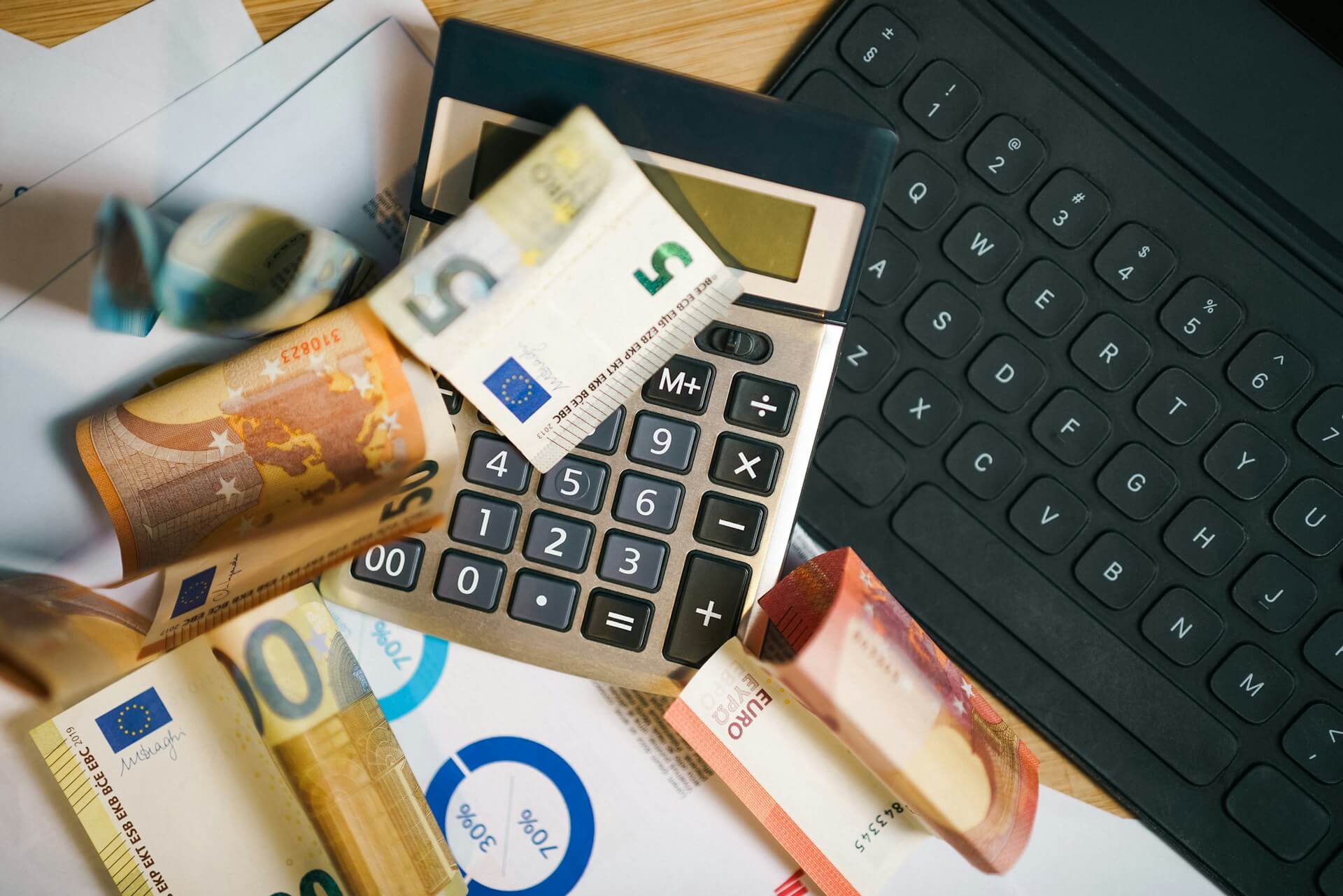 Várias notas de euro espalhadas em cima de uma calculadora, ao lado de um teclado e documentos com gráficos, representando análise financeira ou gestão de despesas.