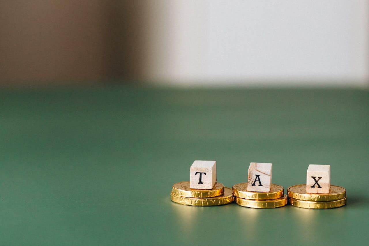 Três pilhas de moedas douradas sobre superfície verde com cubos de madeira que formam a sigla TAX, simbolizando impostos