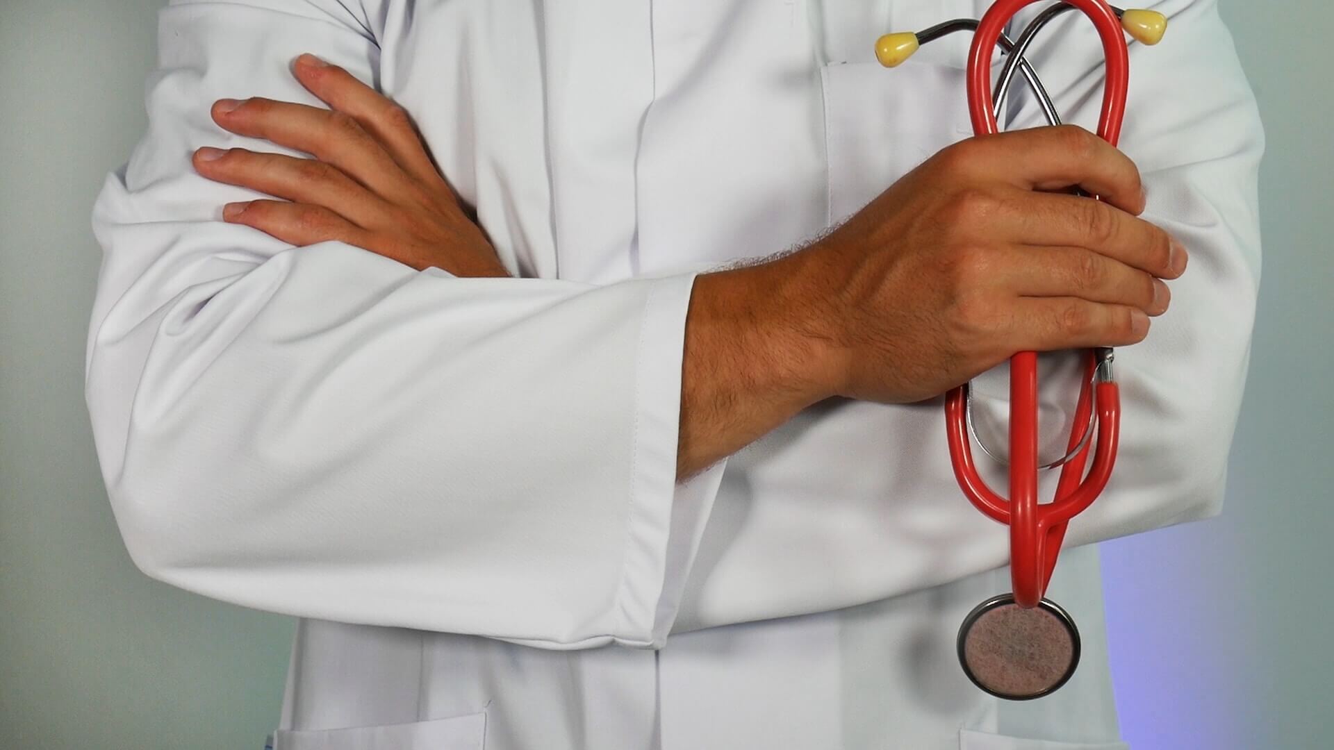 Médico de bata branca com braços cruzados segurando um estetoscópio vermelho