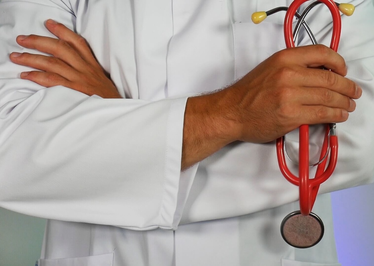 Mãos de médico cruzadas sobre bata branca com estetoscópio vermelho pendurado