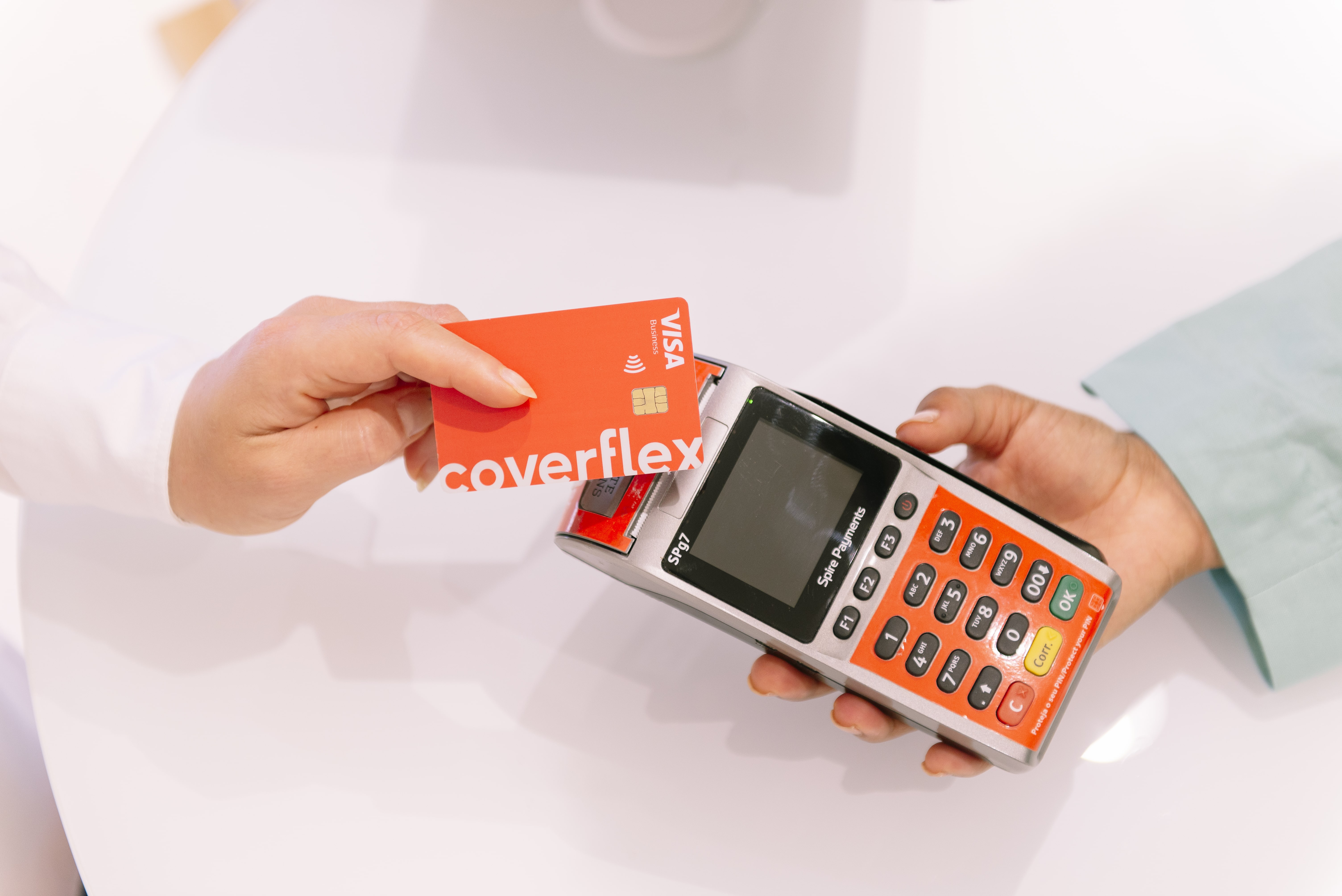 Mão segurando cartão vermelho Coverflex Visa Business junto a terminal de pagamento contactless segurado por outra mão sobre mesa branca