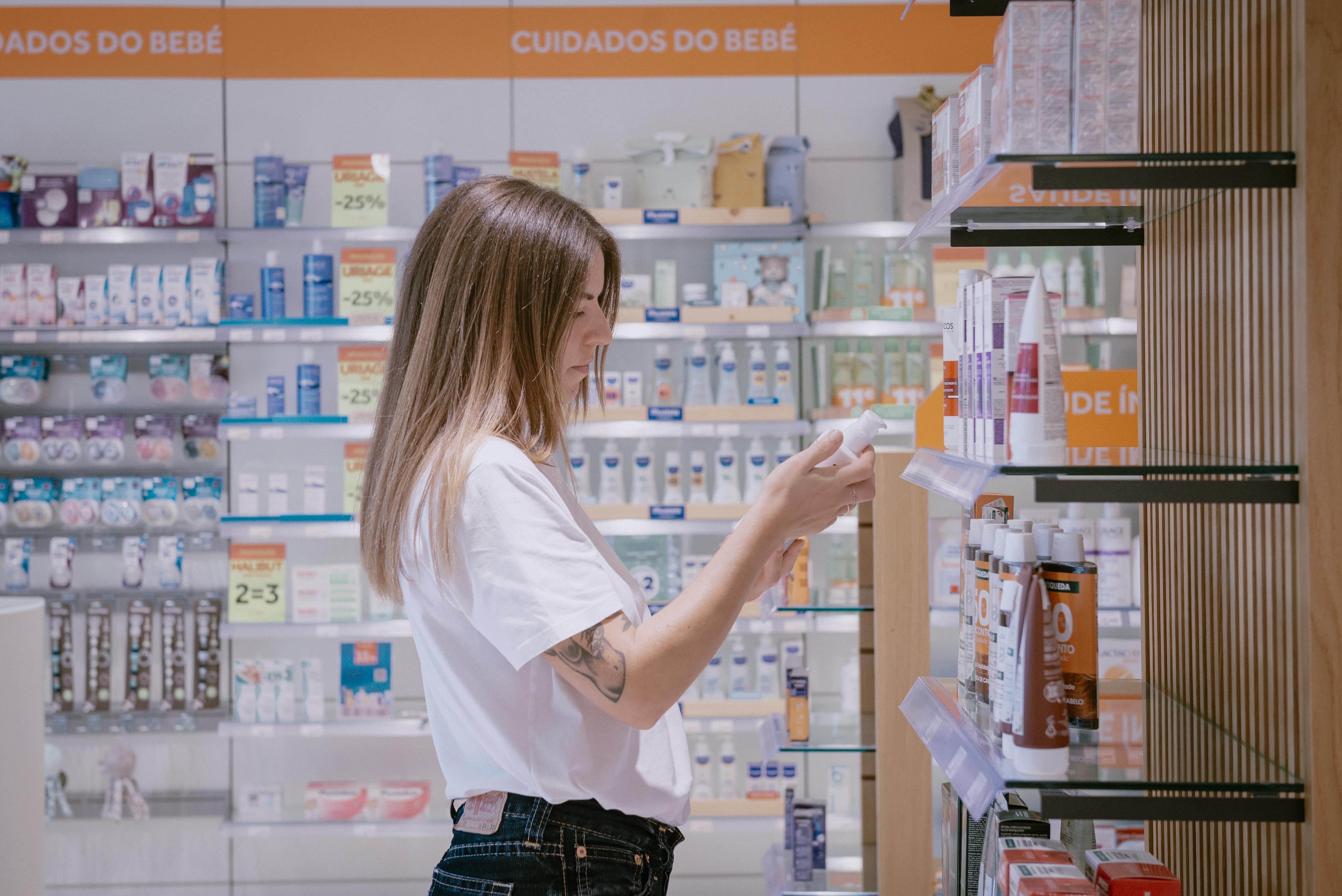 Perfil de jovem mulher com tatuagem no antebraço a examinar frasco branco na secção de cuidados do bebé numa farmácia moderna rodeada de prateleiras com produtos infantis