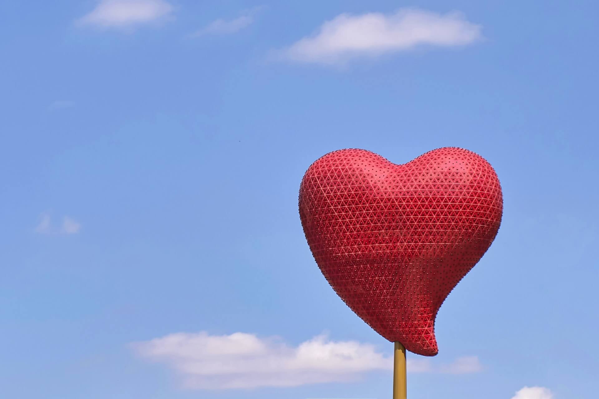 Escultura exterior em forma de coração vermelho com padrão triangular num poste dourado, sobre fundo de céu azul salpicado de nuvens brancas