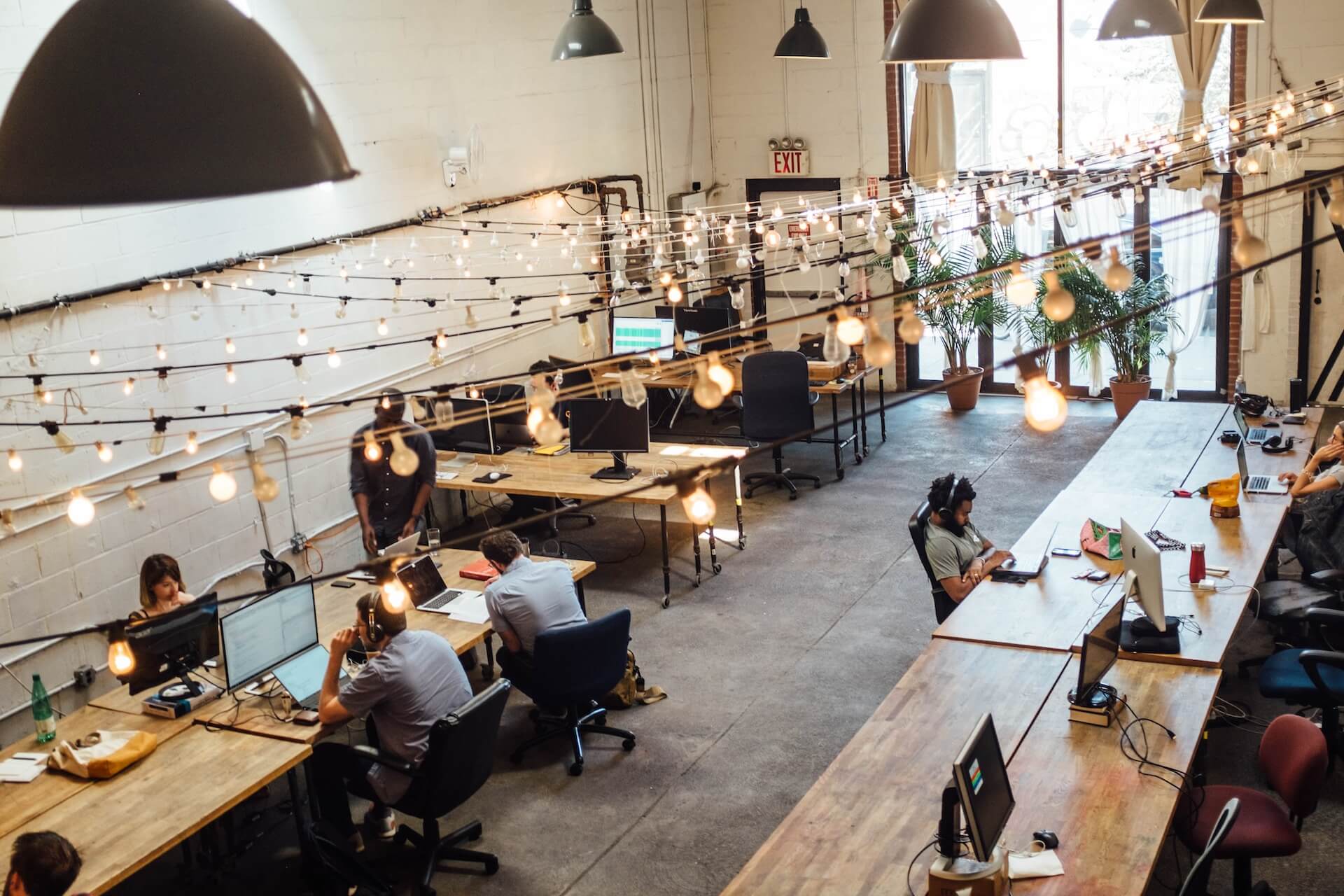 espaco de coworking industrial com iluminacao de lampadas pendentes e varios funcionarios a usar portateis e computadores em mesas de madeira
