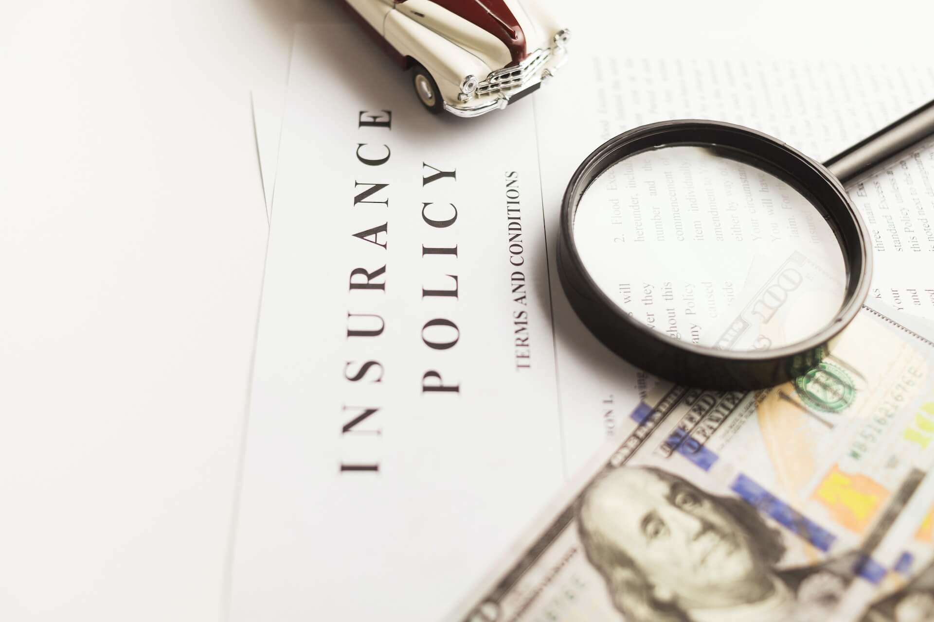 Plano detalhe de carro vintage sobre documento de insurance policy com lupa a destacar termos e condições e notas de cem dólares ao lado representando custos e cobertura