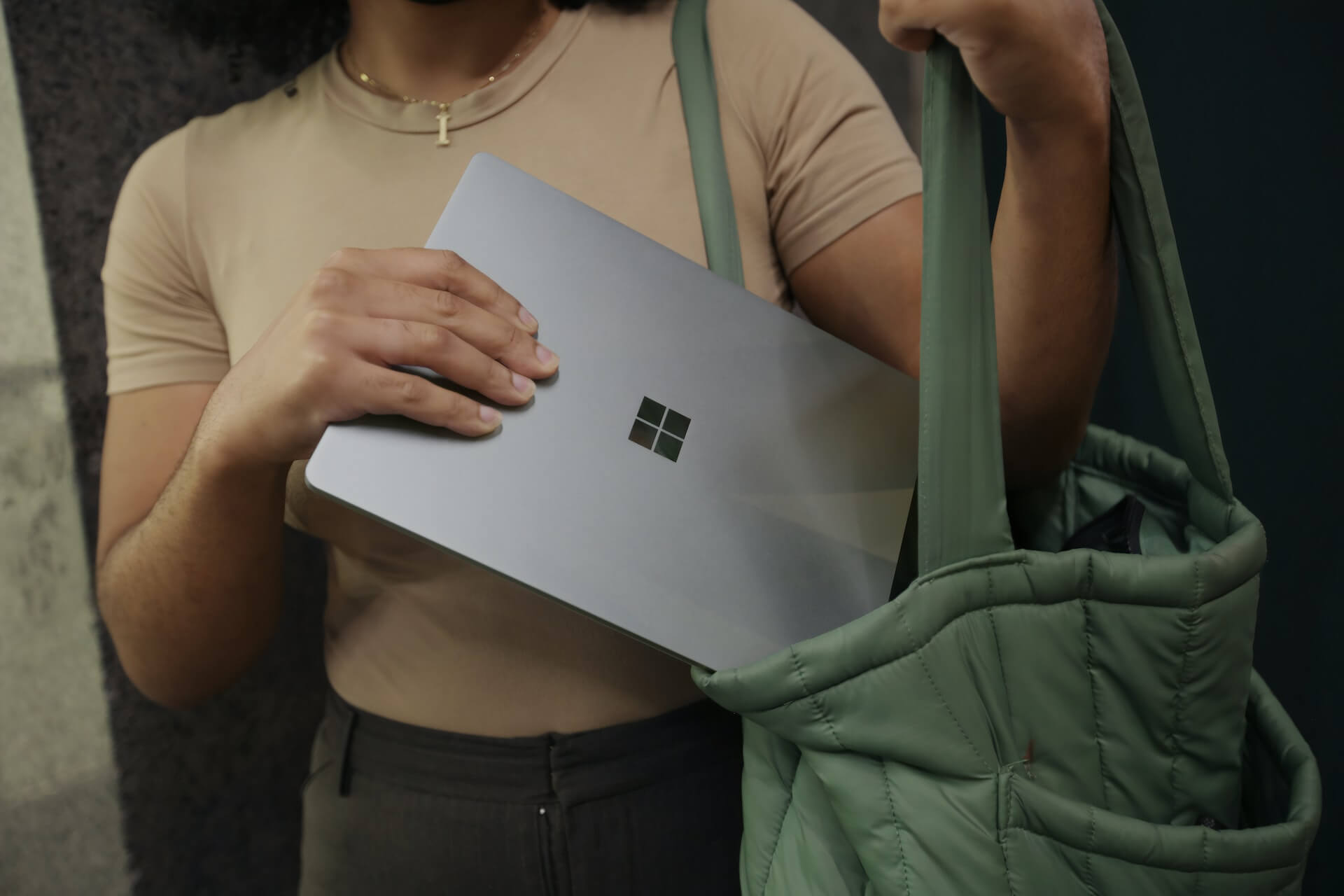mãos de pessoa com t-shirt bege a inserir portátil com logótipo Microsoft numa mala verde acolchoada simbolizando mobilidade e trabalho flexível
