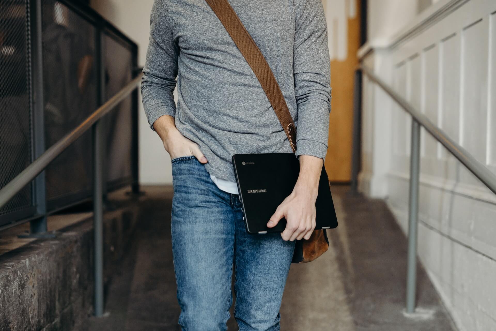 Pessoa vestida com jeans azul e camisola cinzenta segura um portátil Samsung Chromebook debaixo do braço e transporta uma mala de tiracolo castanha enquanto caminha num corredor interior com corrimãos metálicos